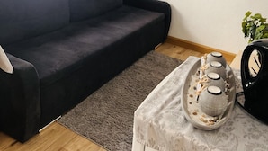 A modern living room with a dark grey sofa, a small table with a decorative item, a view of a cityscape at sunset, and a window with blinds. - Lovely 1-bed Apartment in Koblenz (Koblenz)
