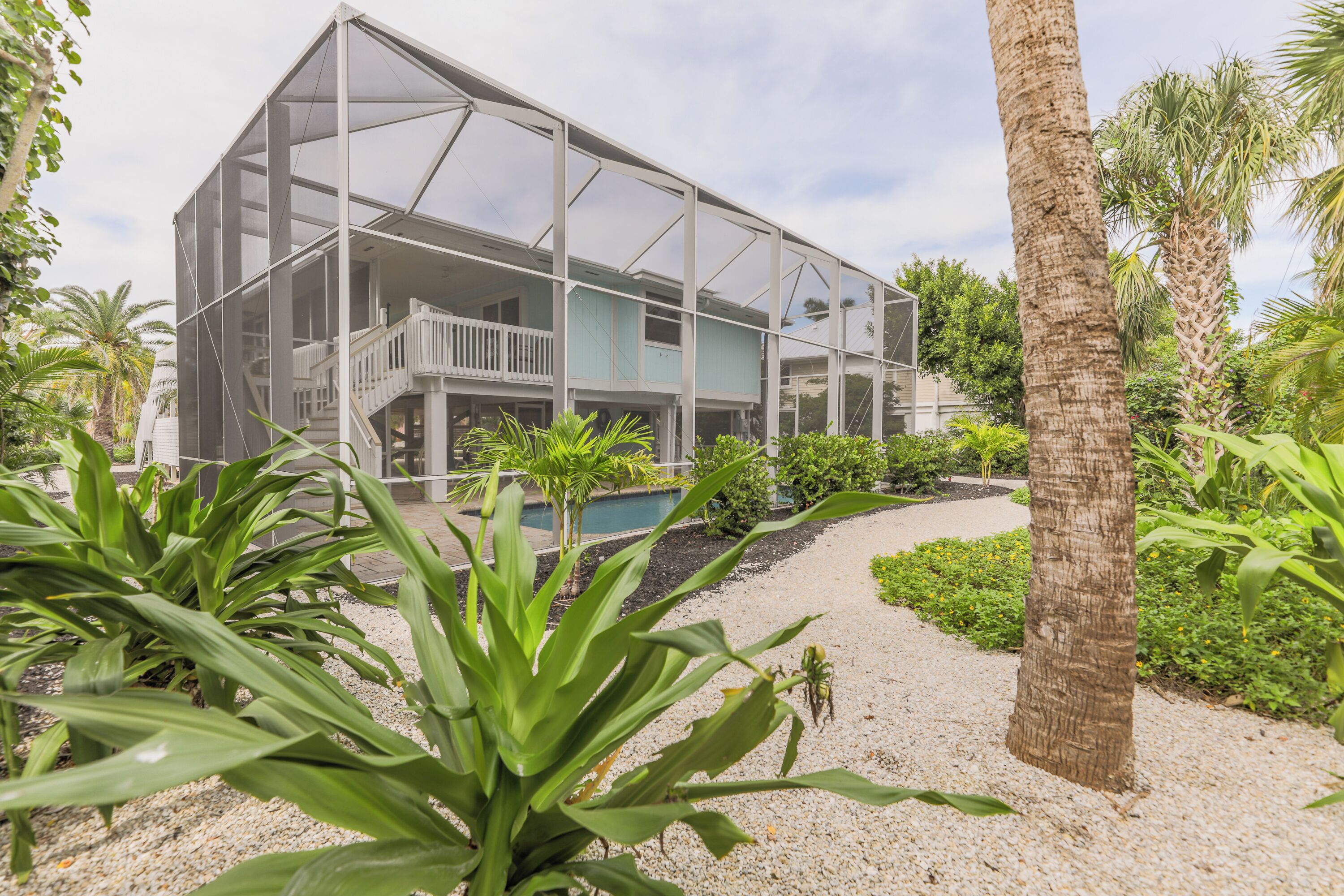 A modern house with a balcony, surrounded by tropical plants and a gravel path.