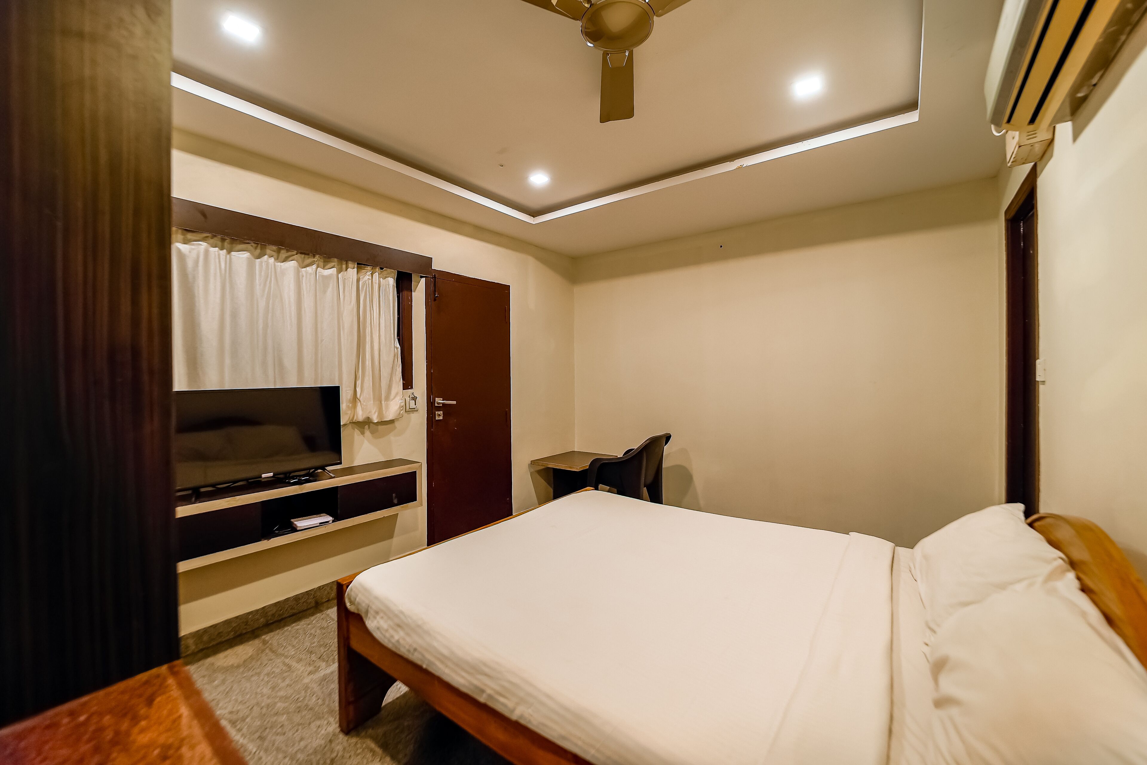 A bedroom with a bed, a TV on a stand, a desk, and a ceiling fan.