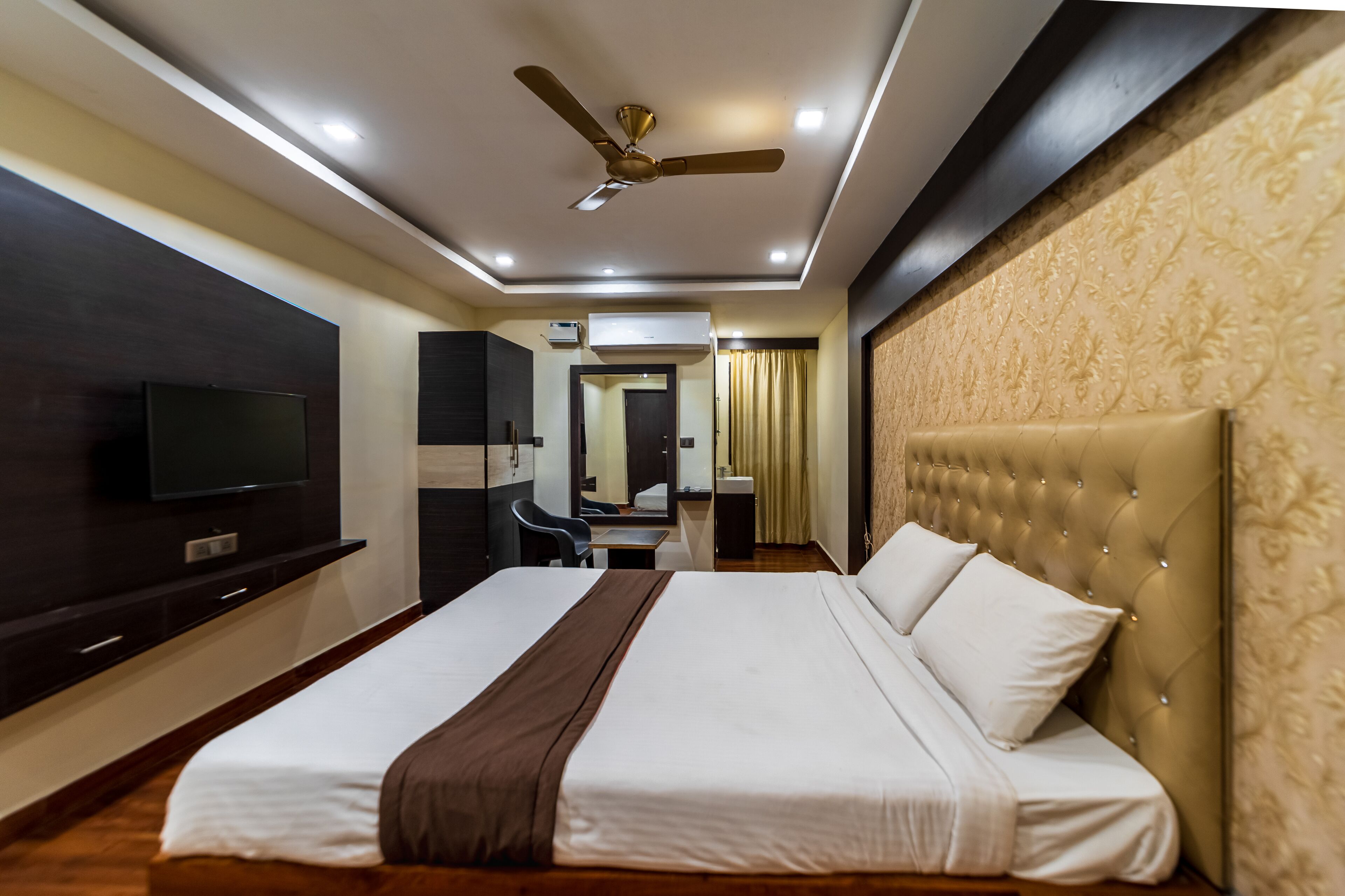 A hotel room with a large bed, a television on the wall, a ceiling fan, and a desk with a chair.