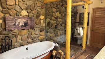 Uma parede de pedra, uma banheira de madeira, um box de vidro e uma porta de madeira.
