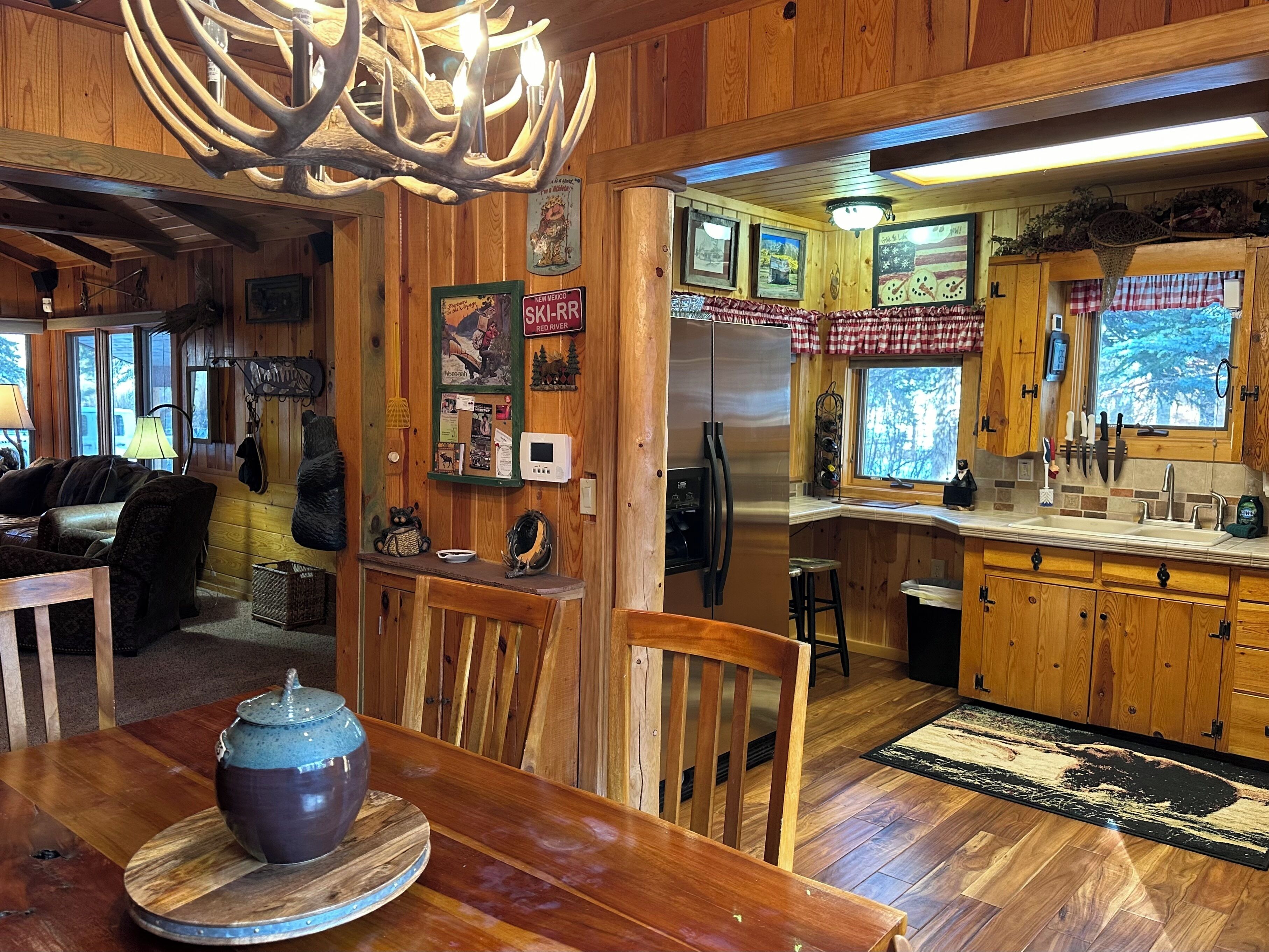 A rustic cabin interior with a wooden dining area, a kitchen with a refrigerator, and a large window overlooking a forest.