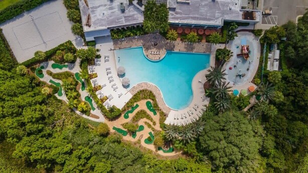 An aerial view of a resort with a large swimming pool, landscaped gardens, and a variety of outdoor seating areas.