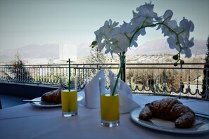 Una mesa con un croissant, un vaso de bebida amarilla y un florero con flores blancas, con vistas a la montaña de fondo.