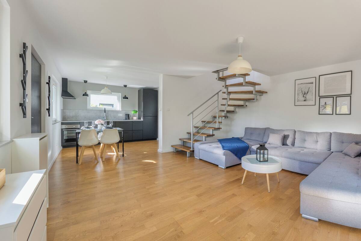 Ein modernes Wohnzimmer mit einem grauen Ecksofa, einem runden Couchtisch und einer Treppe, die zur Küche hinaufführt.