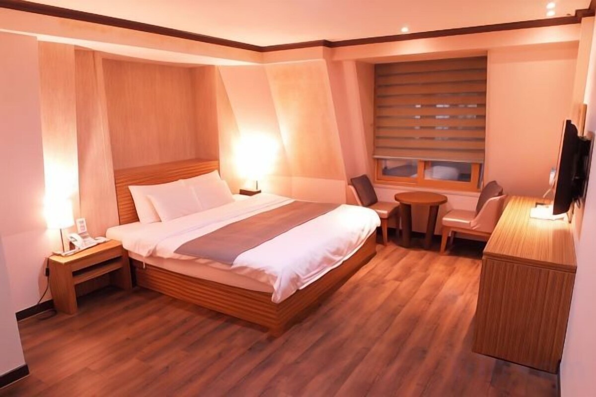 A hotel room with a large bed, bedside tables, a desk, a chair, and a television.