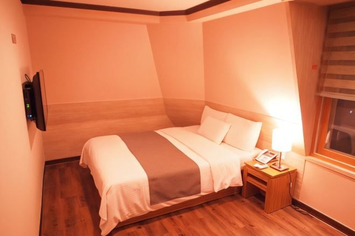 Una habitación de hotel pequeña y acogedora con una cama individual, una mesita de noche con lámpara y una ventana con persianas.