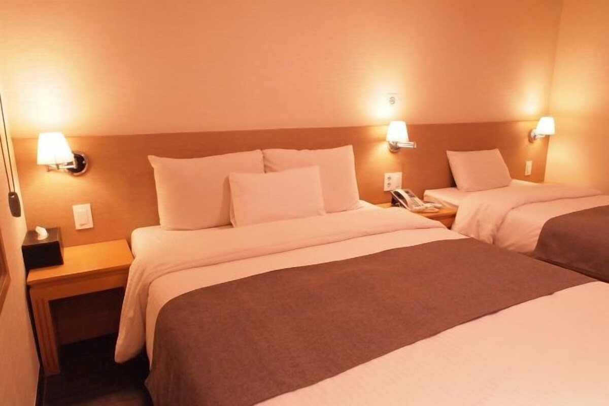 Habitación de hotel con dos camas, cada una con ropa blanca y una manta marrón. Hay lámparas de noche y una mesita con un teléfono y una caja de pañuelos.