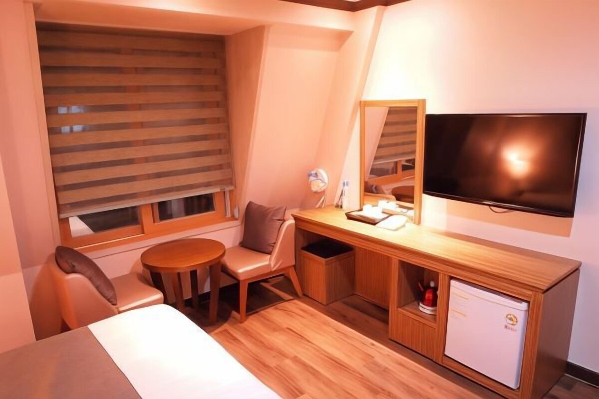Una habitación de hotel pequeña con cama, escritorio, silla, mesita, televisión y refrigerador.