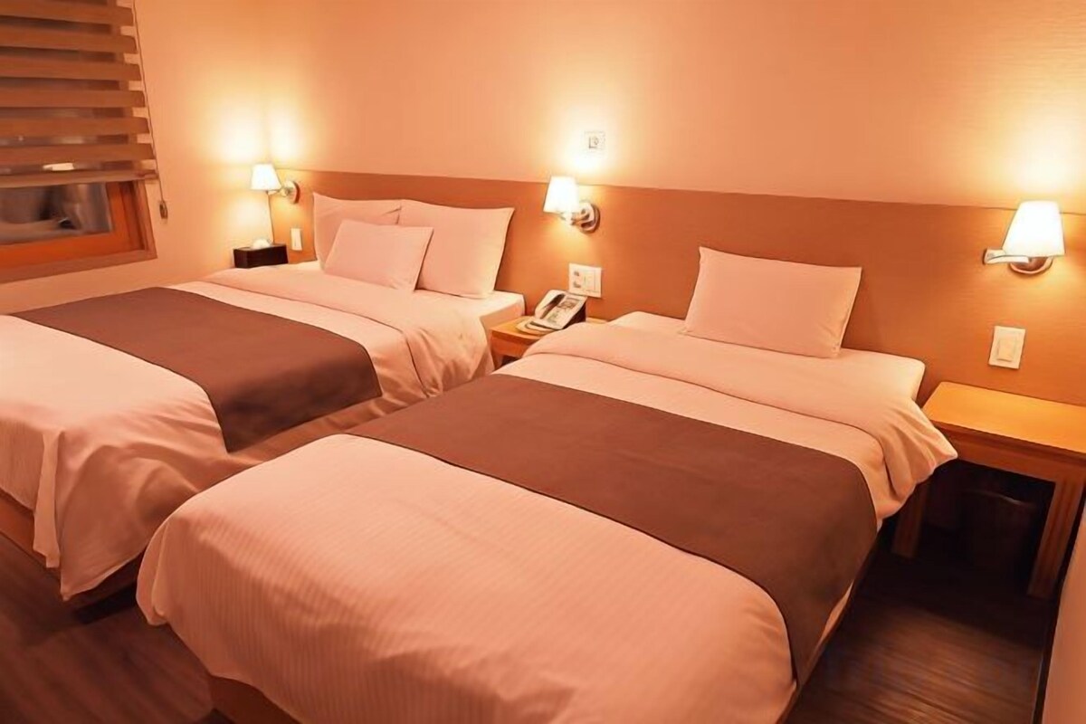 A hotel room with two beds, bedside lamps, a nightstand, and a window with blinds.