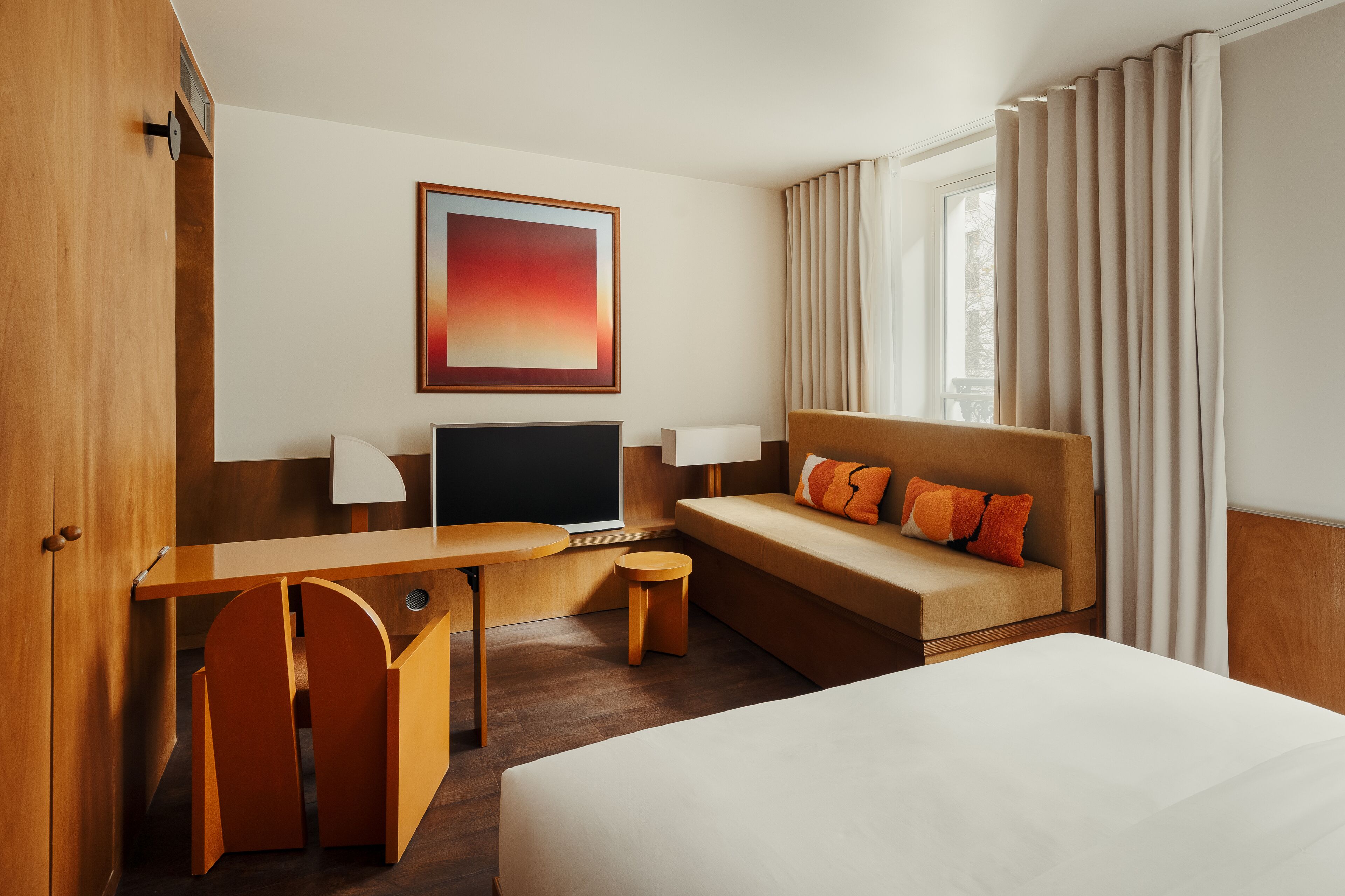 A hotel room with a bed, a desk, a chair, a TV, a sofa, and a window with curtains.