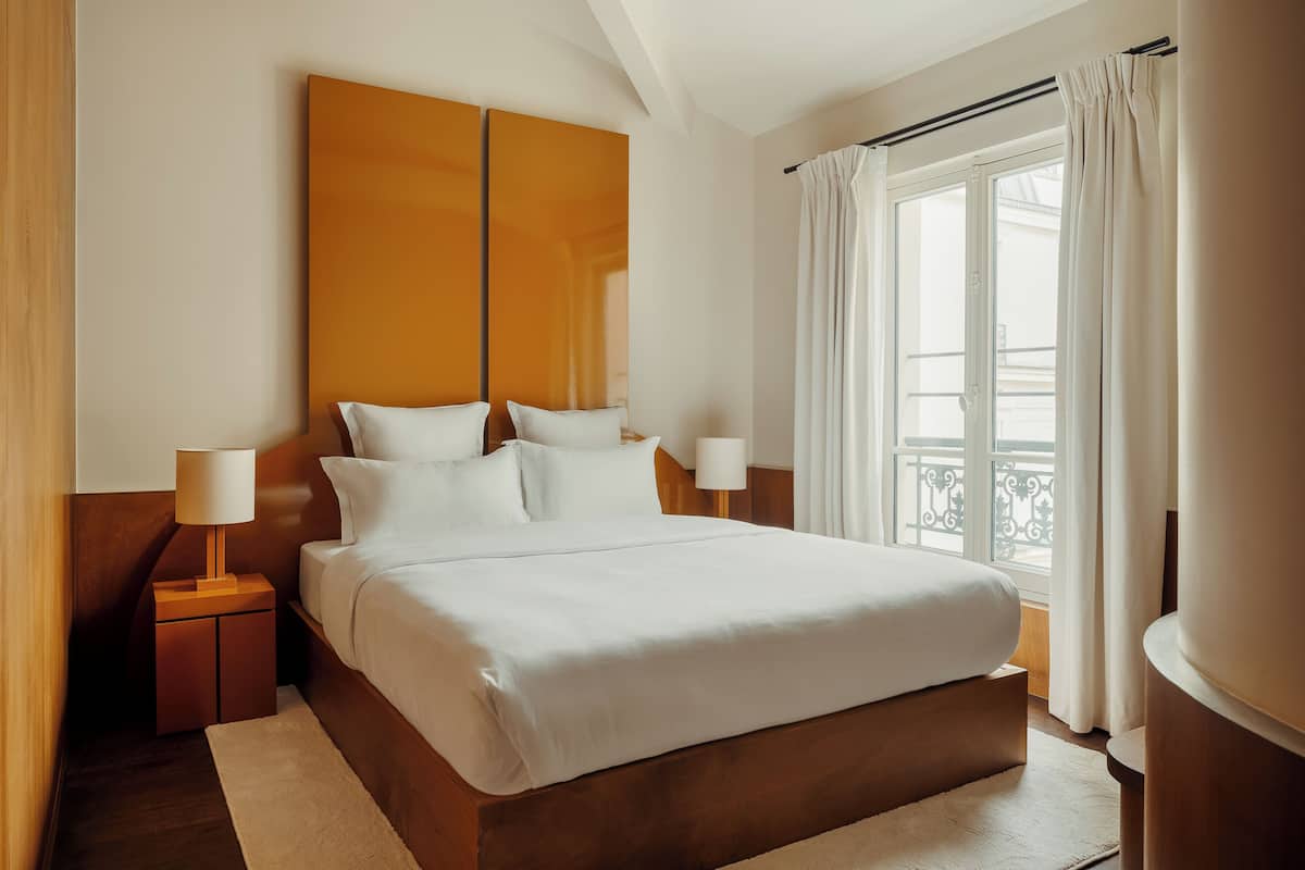 A hotel room with a large bed, two bedside tables, a nightstand, a window with curtains, and a view of the outside balcony.