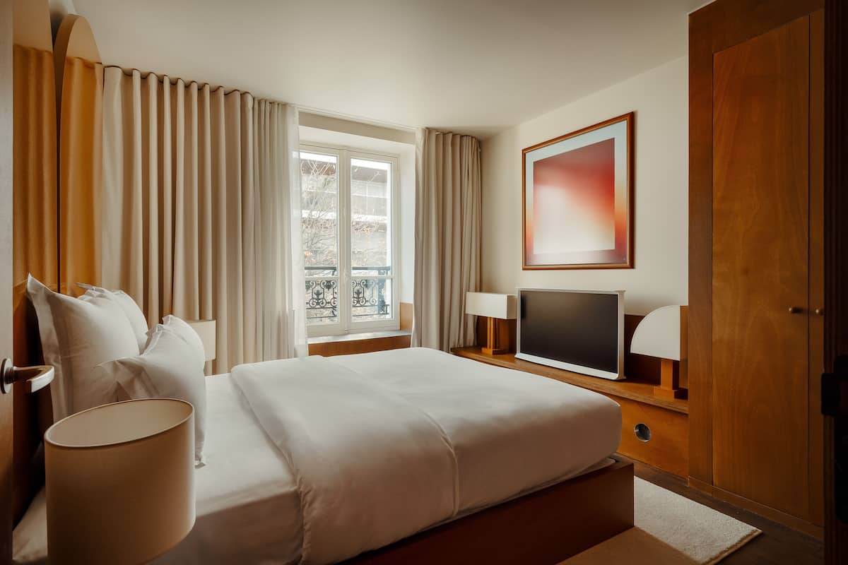 A hotel room with a bed, a TV, a lamp, a window with curtains, and a door.