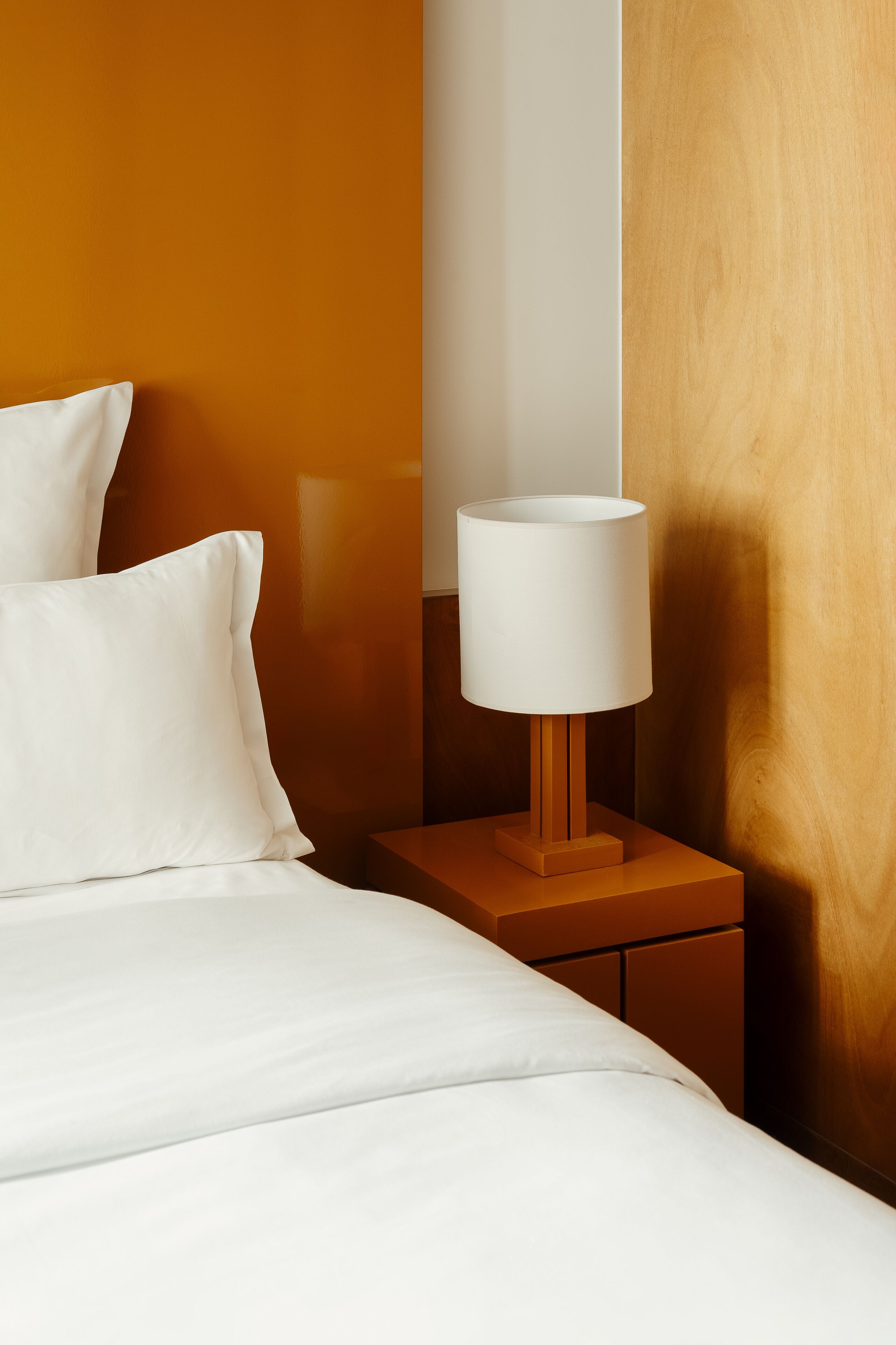 Un lit bien fait, avec du linge de lit blanc, une table de chevet en bois surmontée d’une lampe blanche et un mur en bois.