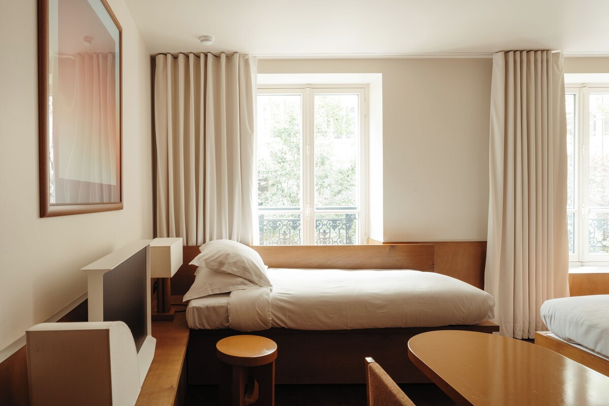 Habitación de hotel con cama, escritorio, silla, ventana con cortinas y vista a zonas verdes.