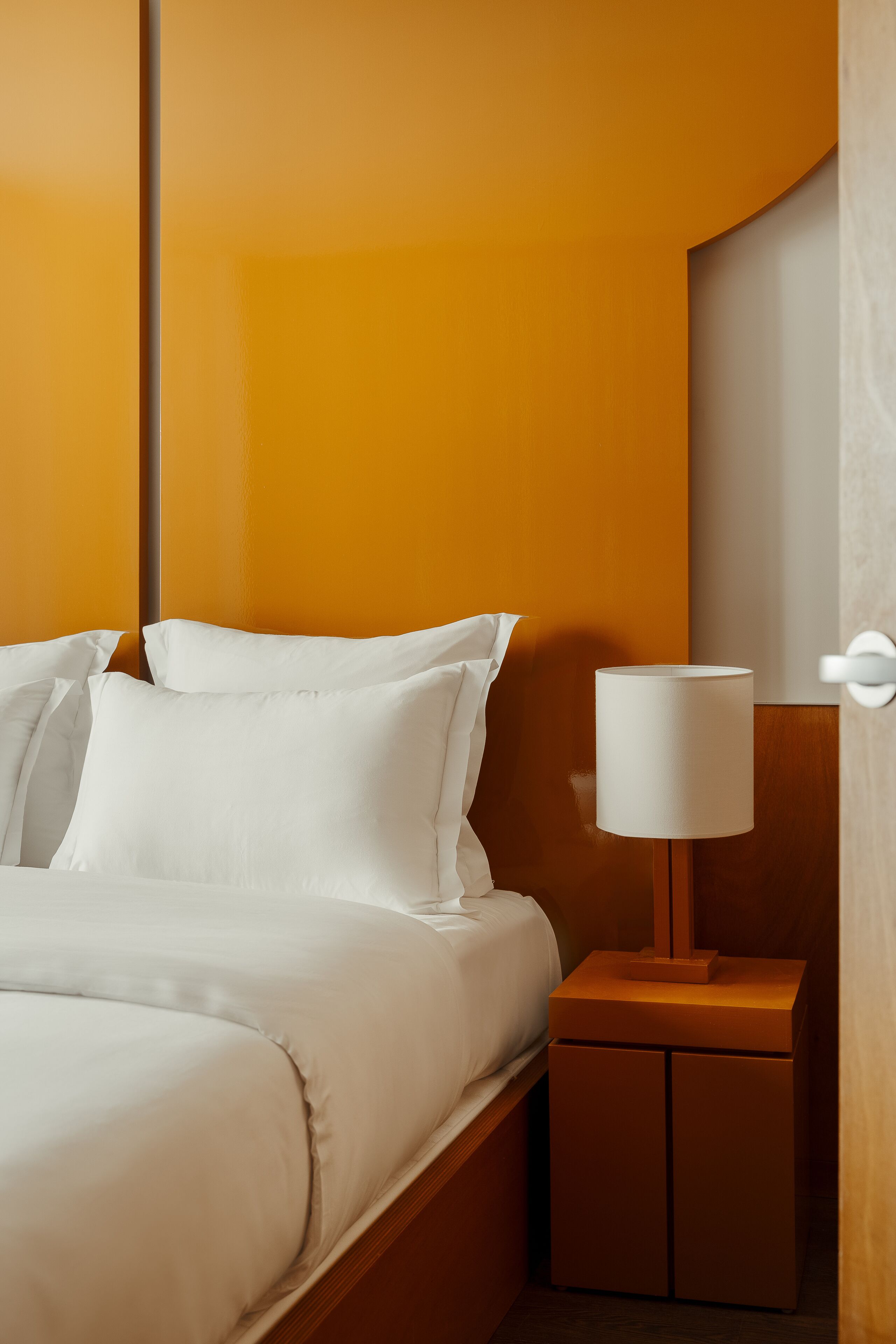 A neatly made bed with white linens, a wooden nightstand with a white lamp, and a wooden headboard.