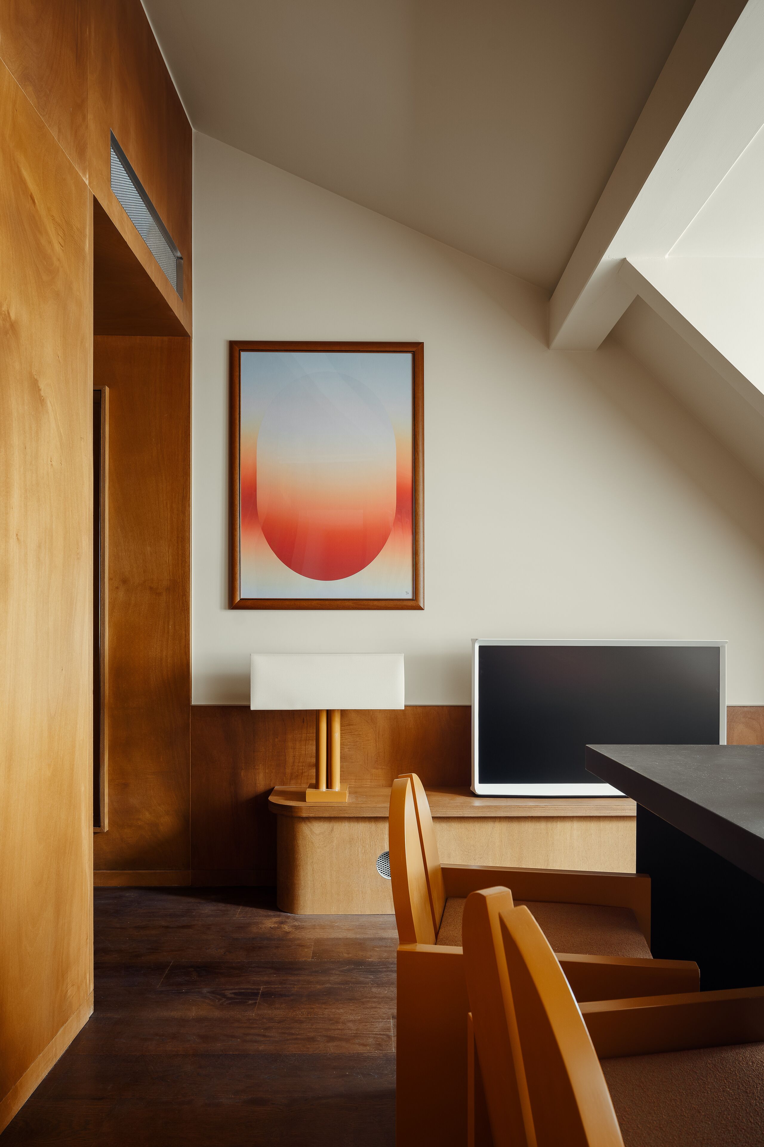 A modern room with a wooden desk, a large framed artwork, and a staircase leading to an upper level.