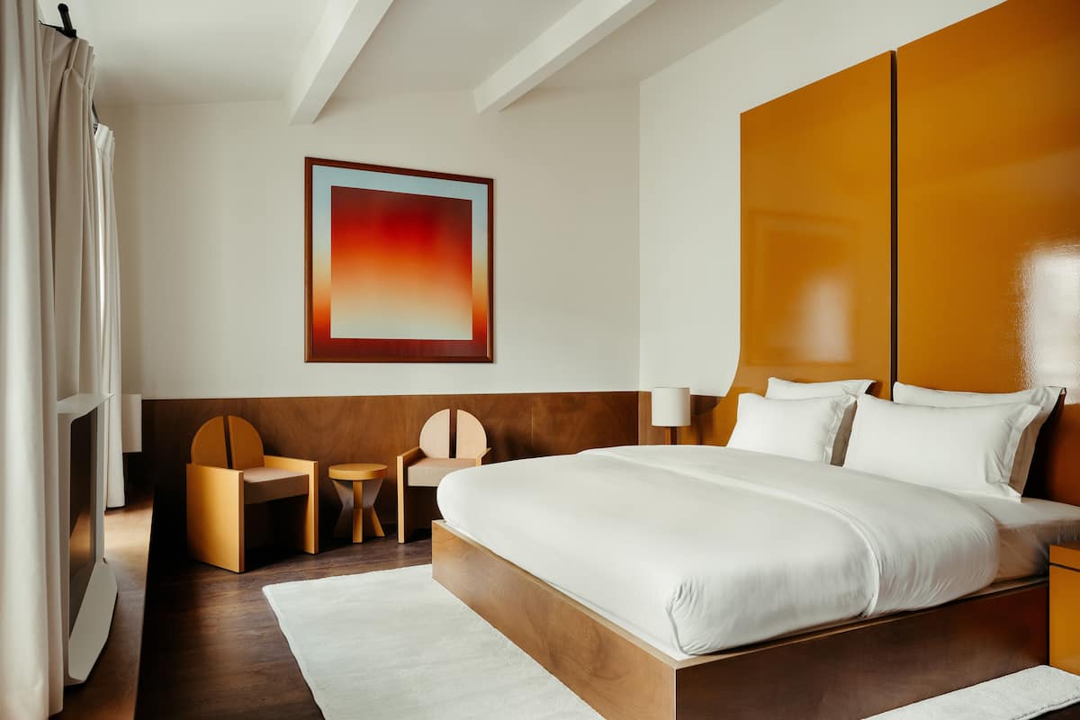 A modern hotel room with a large bed, wooden headboard, a small wooden table, and a chair.