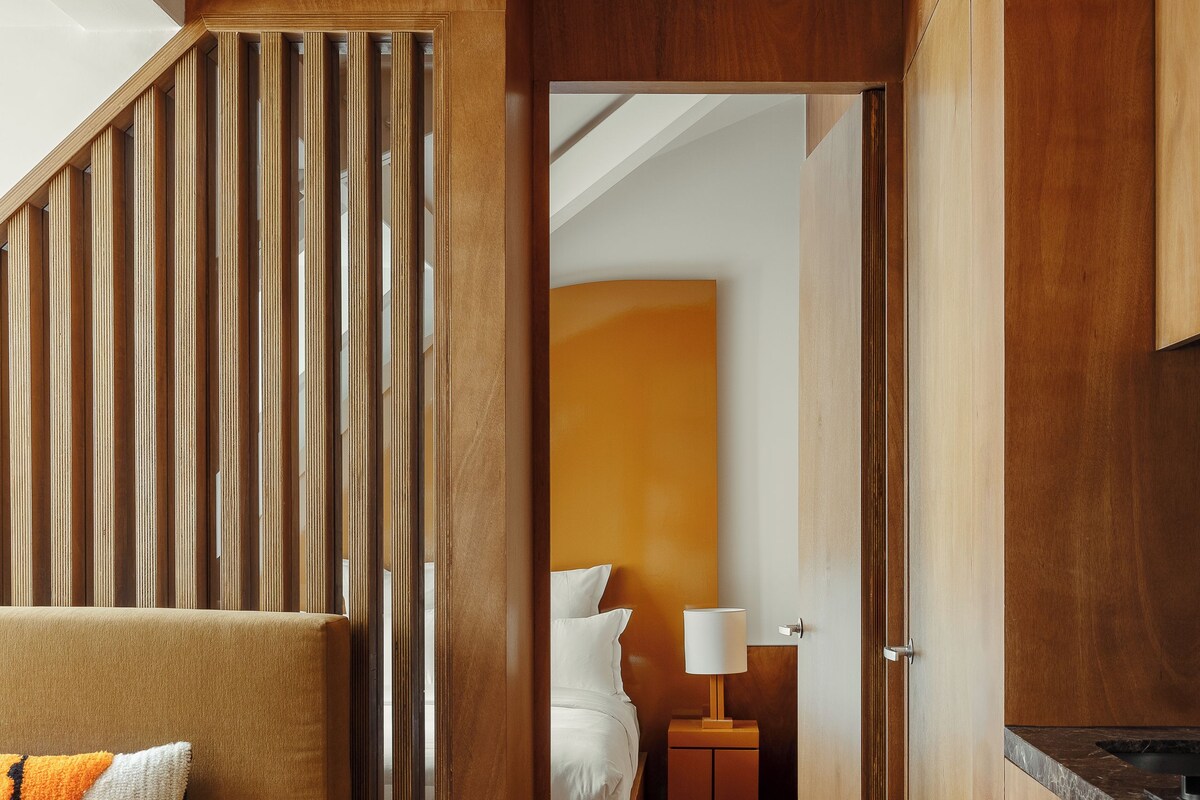 Una habitación de hotel moderna con una escalera de madera, un sofá, una cama, una mesita de noche y una zona de cocina.