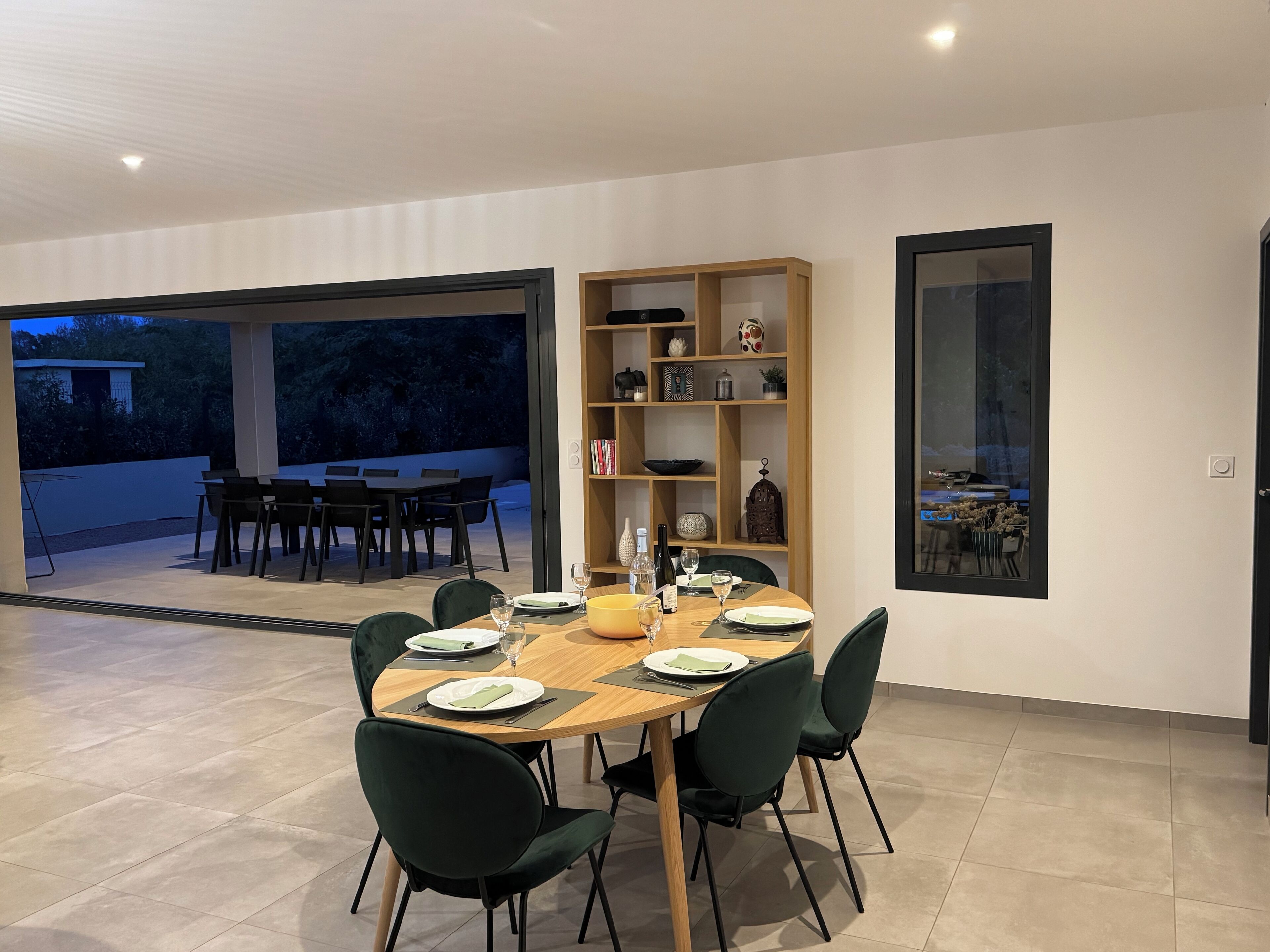 A modern dining area with a wooden table and green chairs, a bookshelf with decorative items, and a view of the outdoors through large glass doors.