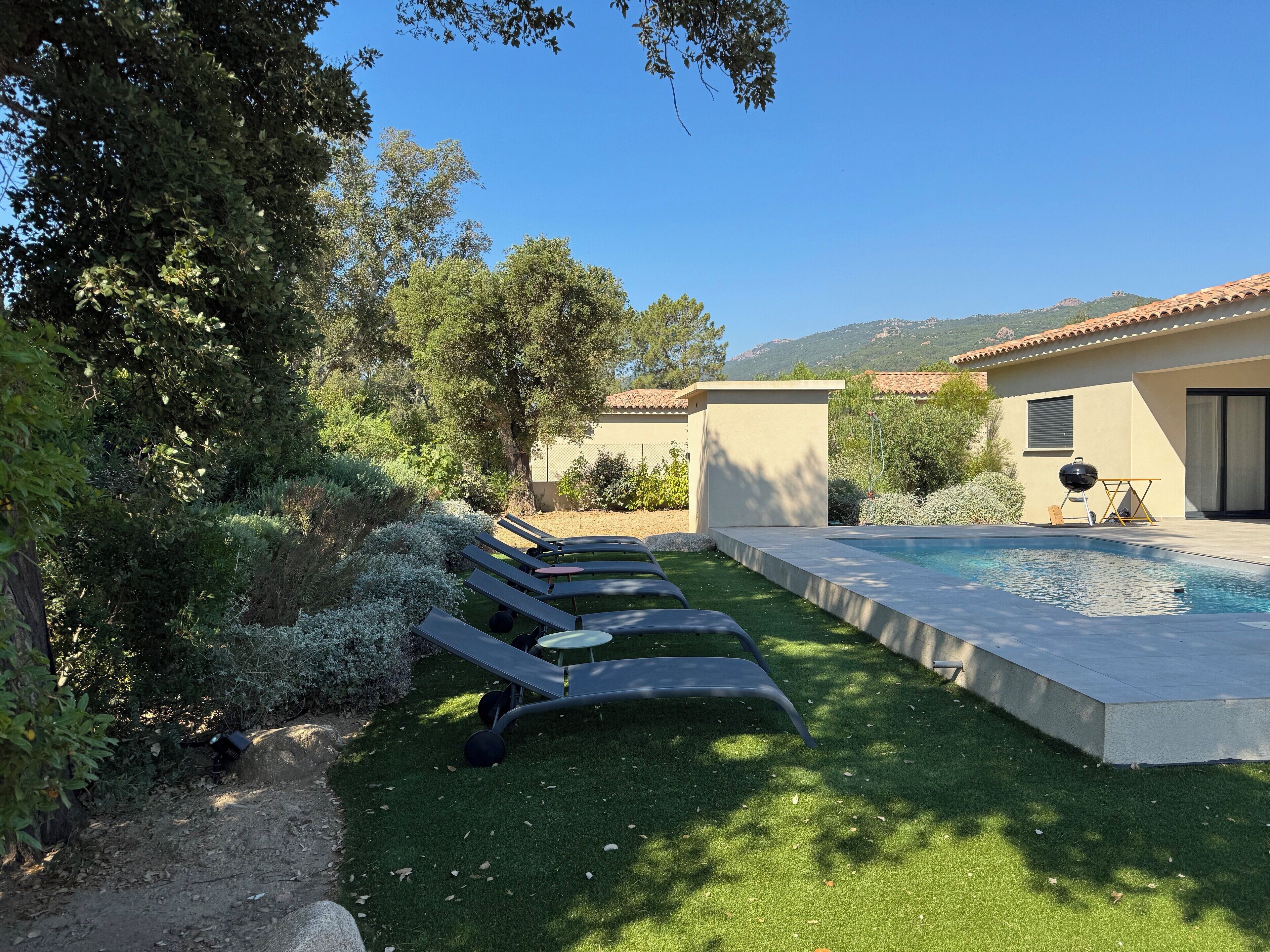 A poolside area with lounge chairs, a barbecue, and a house with a tiled roof.