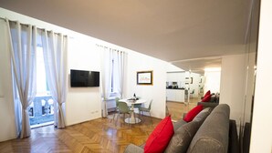 Uma sala de estar moderna com um sofá cinzento, uma mesa de jantar com cadeiras e uma televisão na parede.