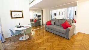 Uma sala de estar moderna com um sofá cinzento, almofadas vermelhas, uma mesa de jantar com cadeiras e um chão de madeira em padrão herringbone.