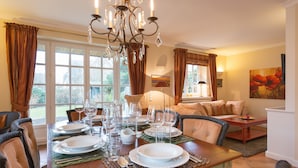 Ein Essbereich mit Tisch für sechs Personen, gedeckt mit Tellern, Gläsern und Besteck. Über dem Tisch hängt eine Chandelier, und der Raum strahlt eine gemütliche Atmosphäre aus, mit einer Couch und einer Lampe im Hintergrund.