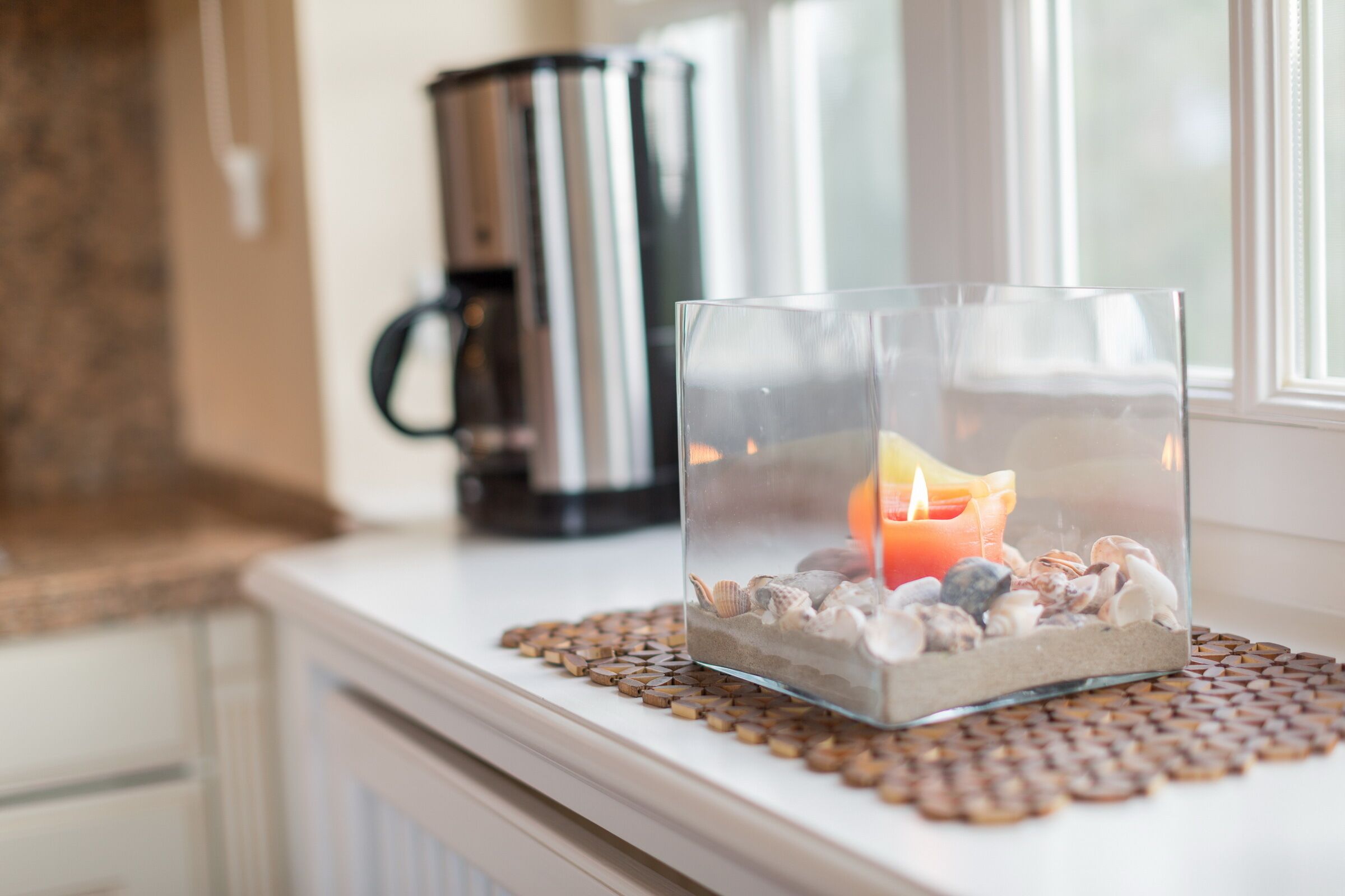 A clear glass candle holder with a lit candle on a textured placemat, placed on a white mantelpiece.
