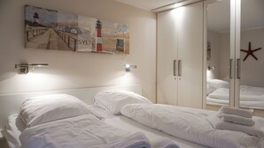 Habitación de hotel con dos camas, ropa de cama blanca, un cuadro de un faro, una decoración con una estrella de mar y un armario con espejo.