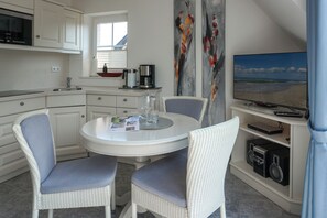 A compact kitchen with white cabinetry, a dining area with two chairs, a coffee maker, and a flat-screen TV displaying a beach scene.