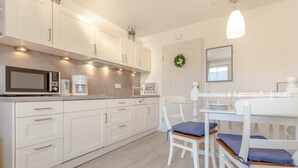 A modern kitchen with white cabinetry, a microwave, and a coffee maker. There is a dining table with chairs and a hanging light fixture. - Abschalten, Erholen, Seelebaumeln - Das Rosenkamp Bietet Entspannung & Erholung (Sylt / OT Westerland)