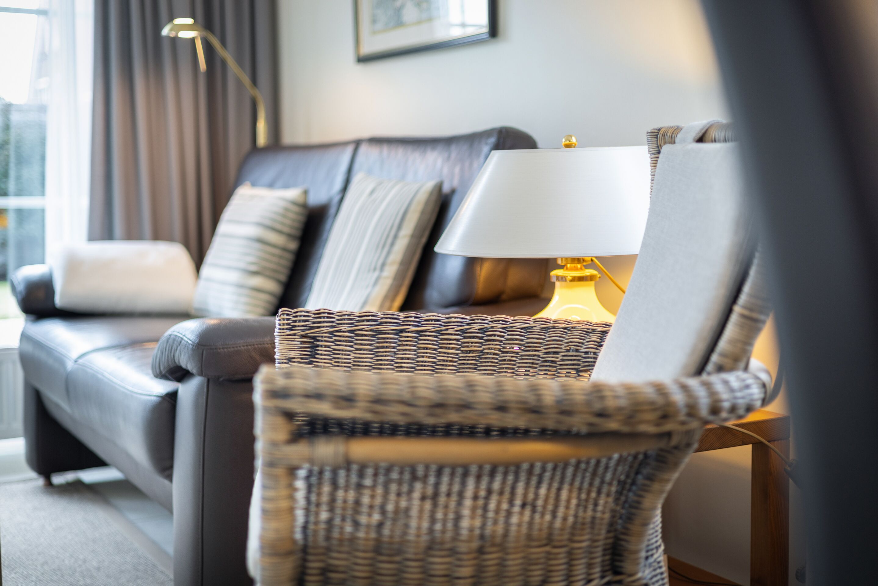 A modern living room with a dark leather sofa, a wicker chair, and a table lamp.