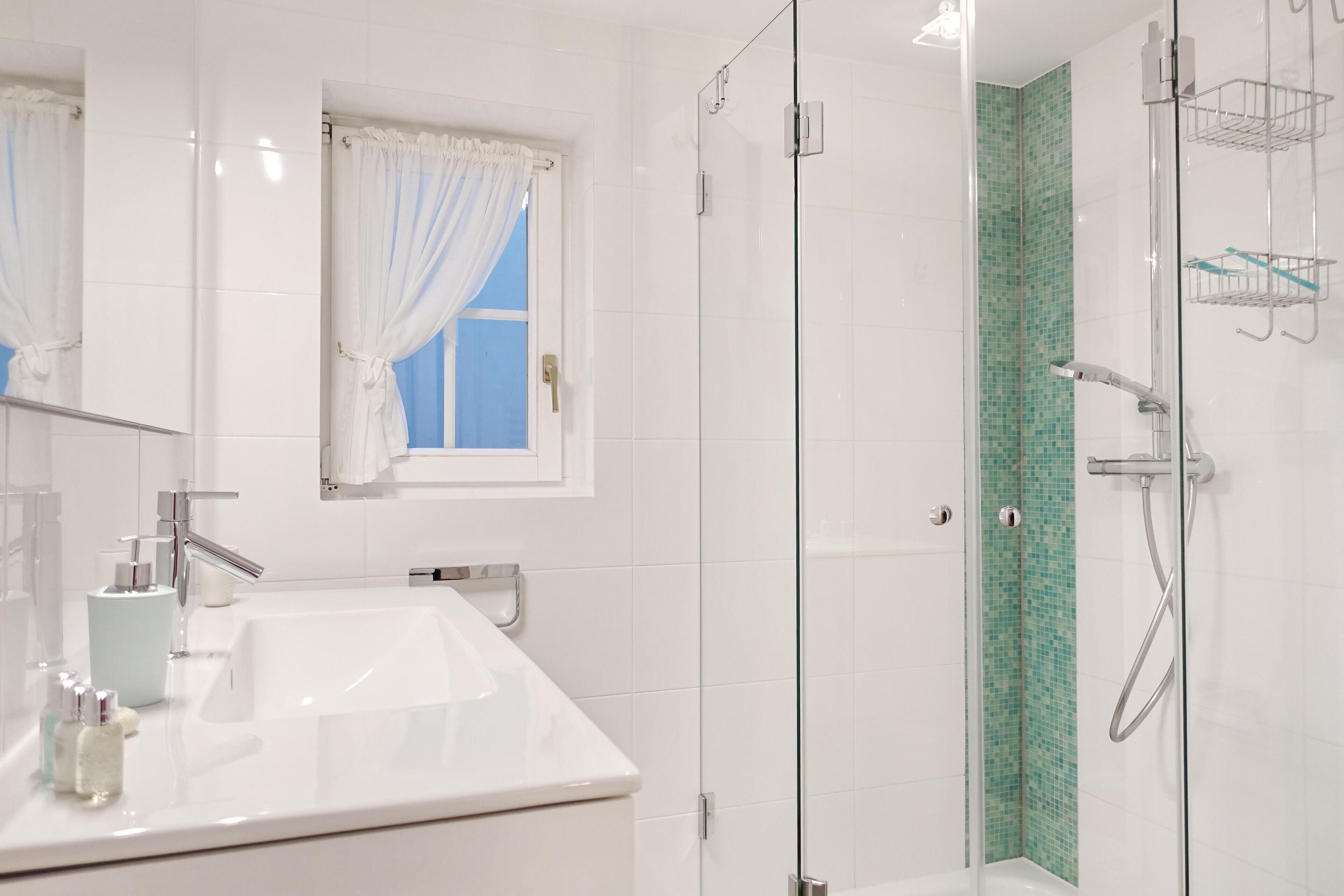 A modern bathroom with a large white sink, a glass shower enclosure, and a small window with white curtains.
