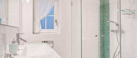 A modern bathroom with a large white sink, a glass shower enclosure, and a small window with white curtains.