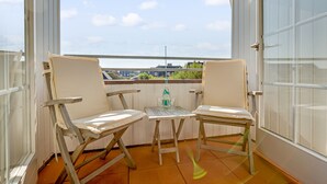 A balcony with two white cushioned chairs and a small table, a bottle of water, and a view of buildings and greenery.