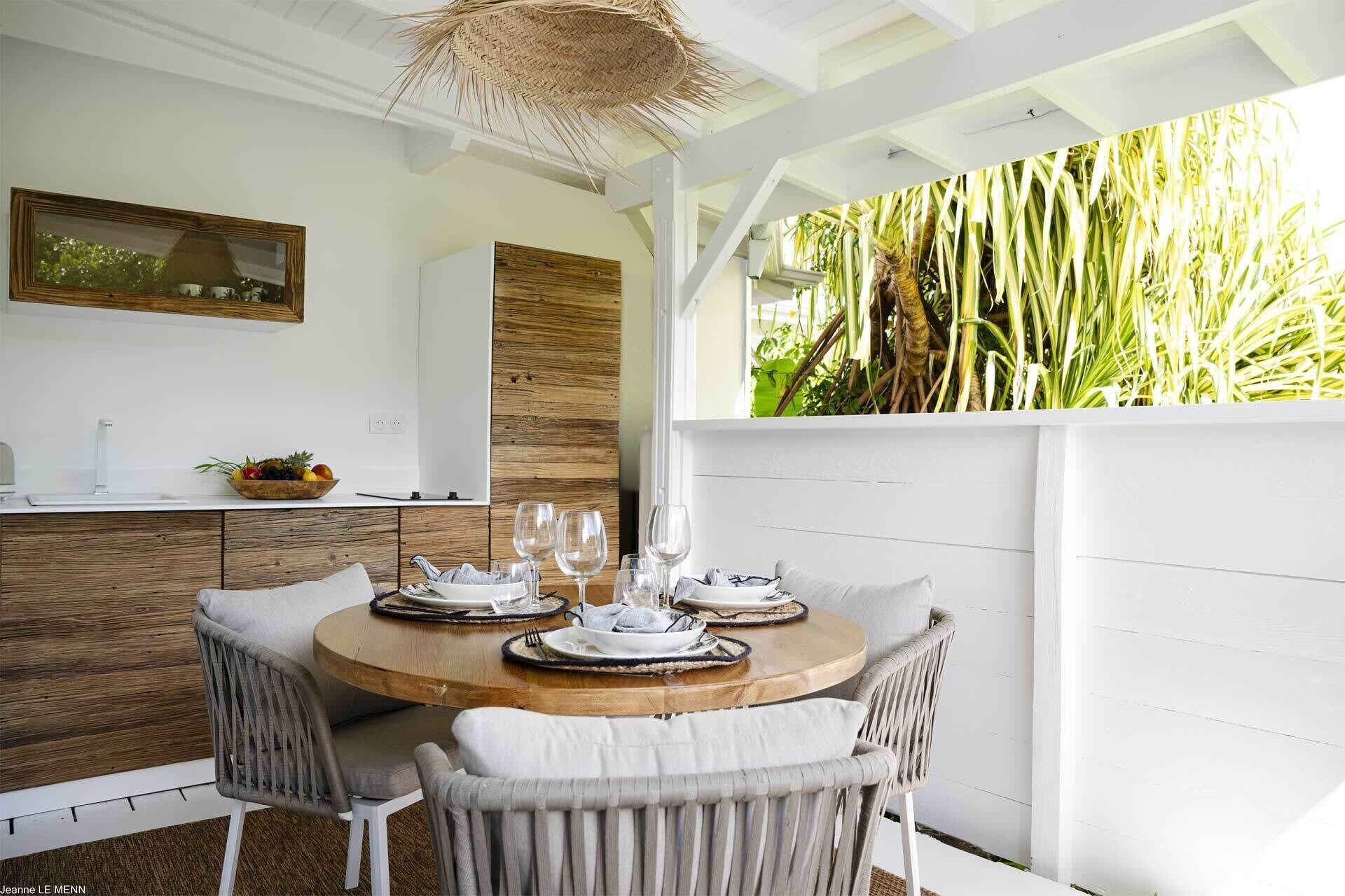 Un comedor con una mesa de madera redonda dispuesta para cuatro personas, rodeada de sillas de mimbre. Se ve una pared blanca con un panel de madera y una lámpara de techo de fibra.
