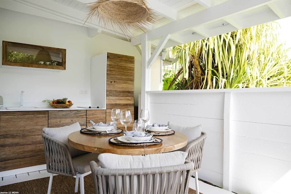 A dining area with a round wooden table set for four, surrounded by wicker chairs. A white wall with a wooden panel and a woven ceiling light fixture is visible. - Pomme Cannelle Luxury Suites & Spa (Orient Bay)