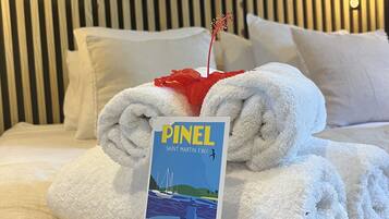 A neatly made bed with white towels, a brochure titled 'PINEL Saint Martin F.W.I.', and decorative wall mirrors.