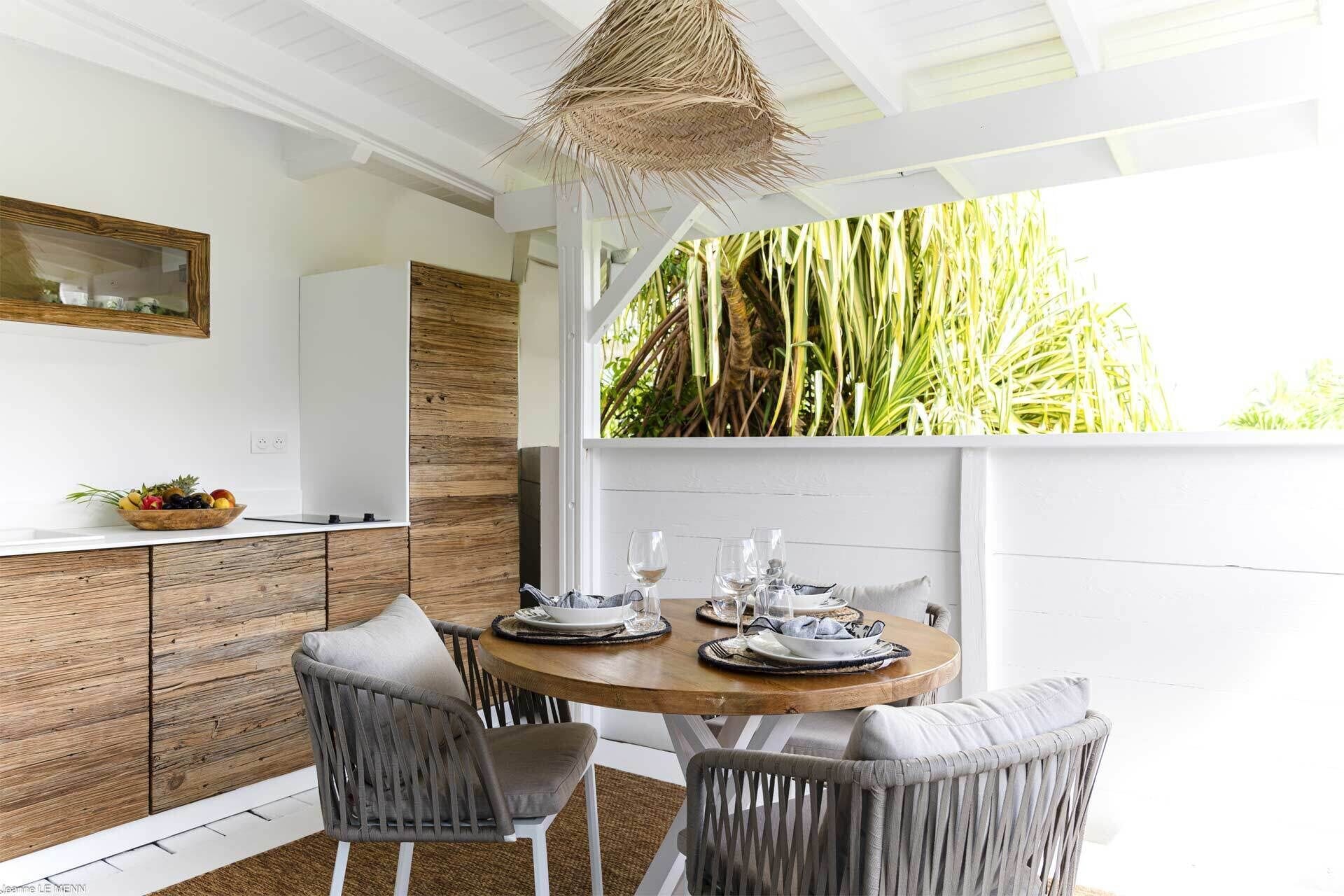 A dining area with a round wooden table set for four, surrounded by wicker chairs. A woven ceiling light hangs above, and a window with a view of tall plants is visible.
