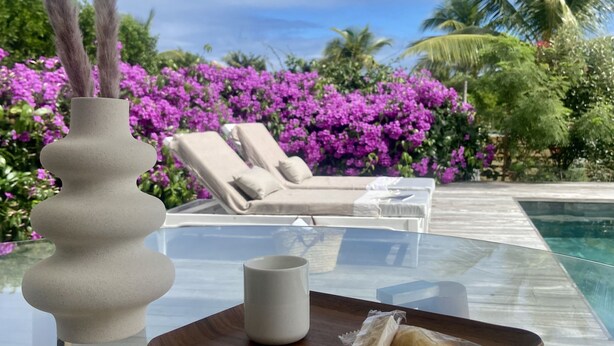 Uma mesa de vidro com uma caneca branca e uma gaveta de pão, um vaso decorativo, e uma área de lounge à beira da piscina com vista para flores roxas e palmeiras.