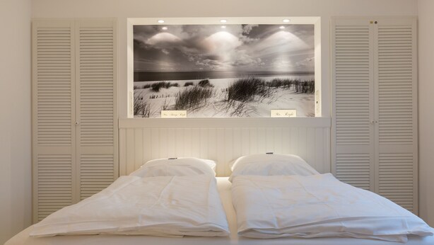 A bedroom with a bed, white bedding, and a large framed picture of a beach scene on the wall. - Feinster Sylt-luxus - Nantucket-style (Wenningstedt-Braderup (Sylt))