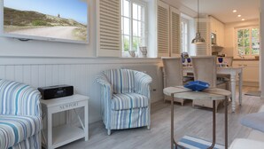 A room with a television displaying a scenic view, a striped armchair, a small table with a radio, and a dining area with a chair and table. - Feinster Sylt-luxus - Nantucket-style (Wenningstedt-Braderup (Sylt))