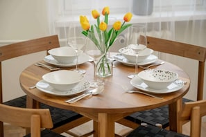 Une table de salle à manger ronde en bois, mise avec des bols blancs, des verres à vin transparents et un vase contenant des tulipes jaunes.
