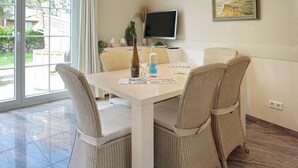 Un coin repas avec une table blanche, des chaises en osier, une bouteille de vin et une bouteille d’eau.