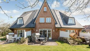 Une maison à deux étages avec une façade en briques, un toit à pignon et une véranda à l’avant, aménagée avec du mobilier d’extérieur.