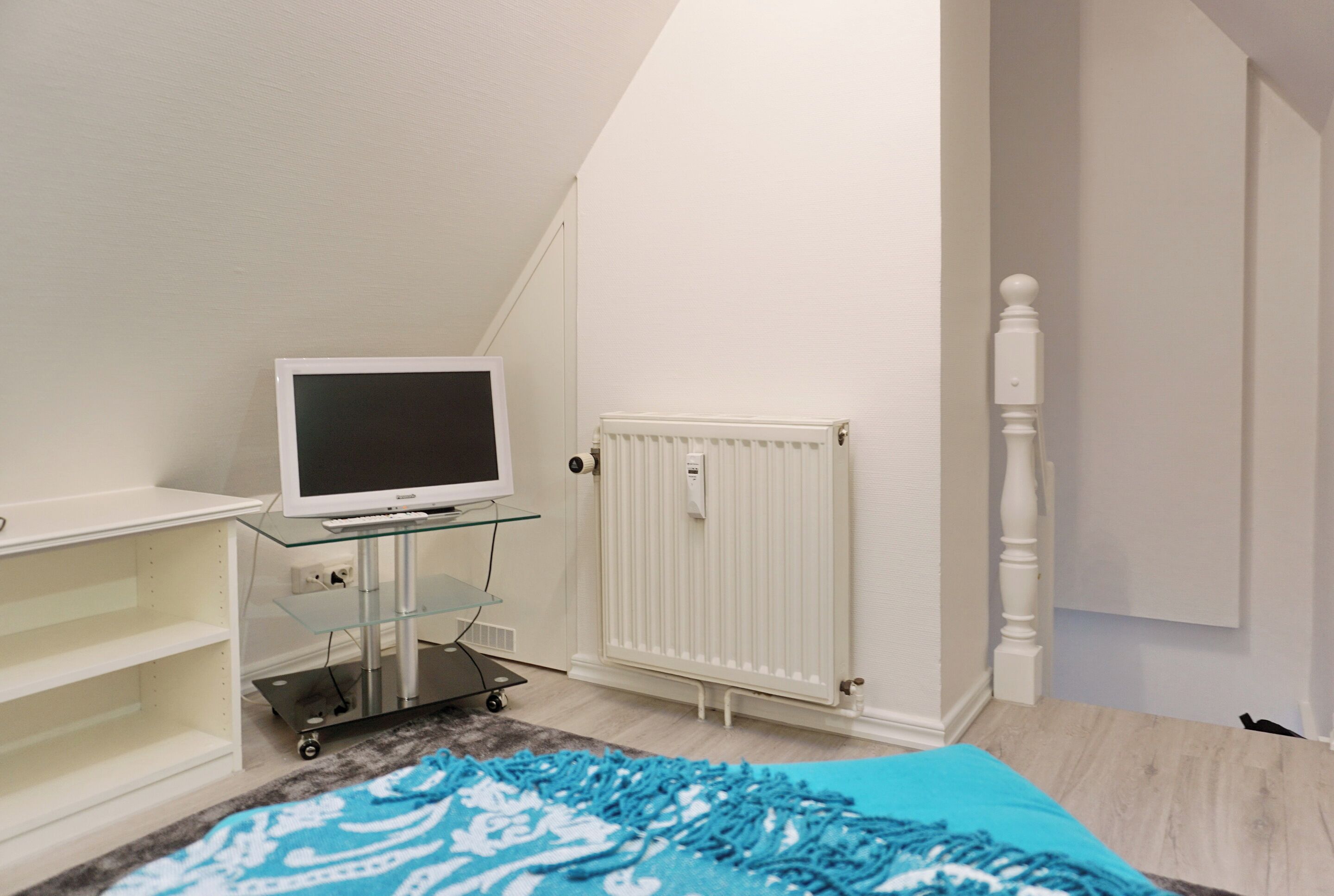 A room with a television on a glass shelf, a radiator, and a bed with a blue and white patterned blanket.