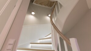 Una escalera con peldaños de madera y paredes blancas, que lleva a un techo con iluminación empotrada.