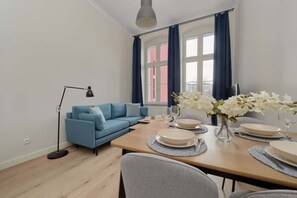 A modern living room with a blue sofa, a dining table set with plates and glasses, a floor lamp, and a window with curtains.