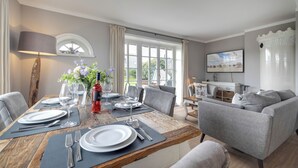 A dining area with a wooden table set for four, surrounded by grey upholstered chairs. A large window with white curtains provides natural light, and a television is mounted on the wall. - Lifestyle Unter Reet, Strand & Meer um die Ecke (Wenningstedt-Braderup (Sylt))
